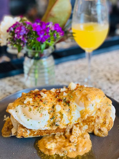 A plate of eggs Benedict topped with creamy sauce, served with a mimosa and a small vase of flowers in the background.