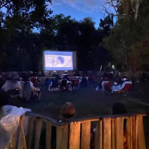 Outdoor movie screening at dusk, with an audience seated on the grass, watching a film on a large screen.