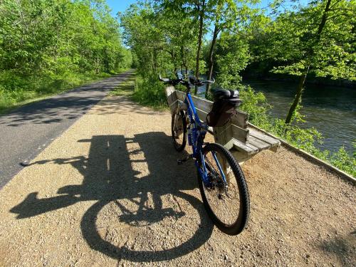 A blue bicycle rests on a path beside a river, with a bench and lush greenery in the background.