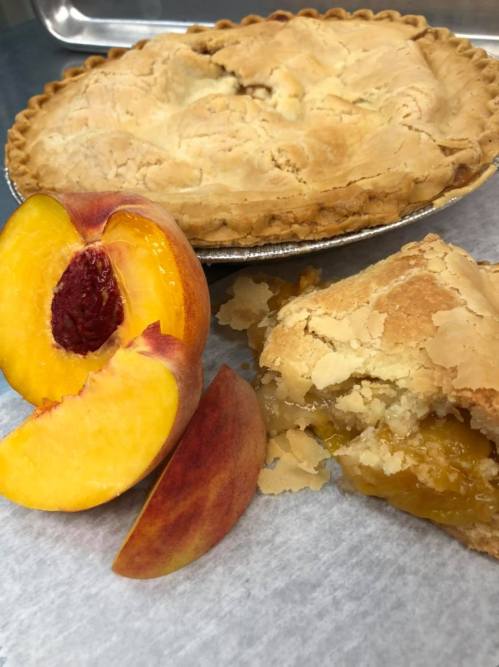 A freshly baked peach pie with a slice cut out, accompanied by a halved peach on a baking sheet.