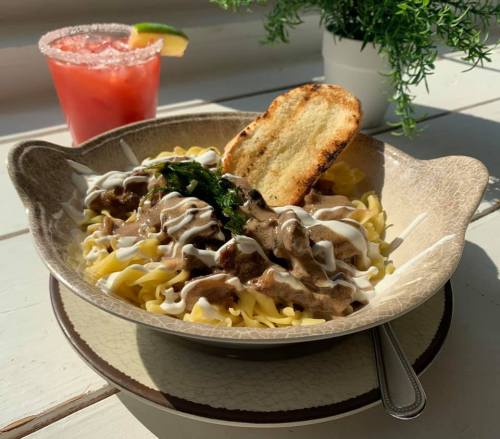 A bowl of pasta topped with creamy sauce and herbs, accompanied by a toasted slice and a colorful drink.