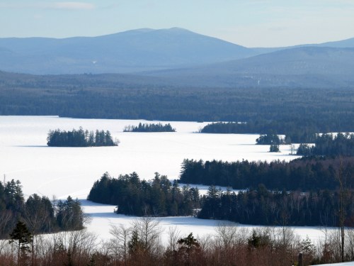 A serene winter landscape featuring a frozen lake surrounded by snow-covered trees and distant mountains.