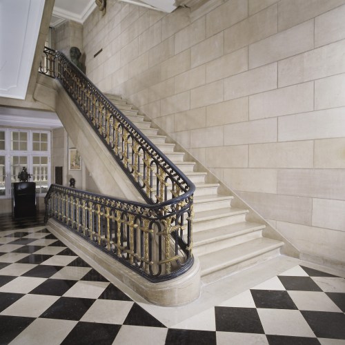 A grand staircase with a decorative black railing, set against a stone wall and a checkered floor.