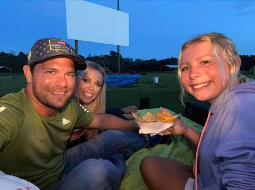 A group of three people enjoying snacks outdoors at dusk, sitting on a blanket with a movie screen in the background.