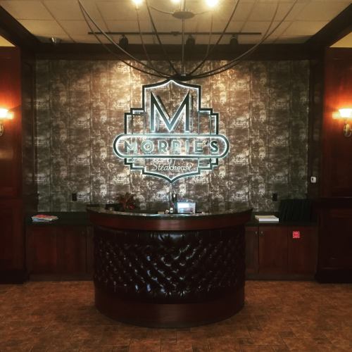 Interior of Morrie's Steakhouse featuring a stylish reception desk and a prominent illuminated logo on the wall.