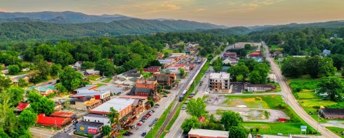 Aerial view of a small town surrounded by mountains, featuring shops, greenery, and a winding road.