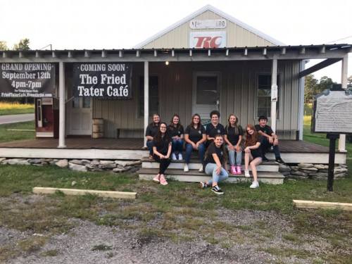 A group of young people poses on the steps of a building with a sign announcing the grand opening of The Fried Tater Café.