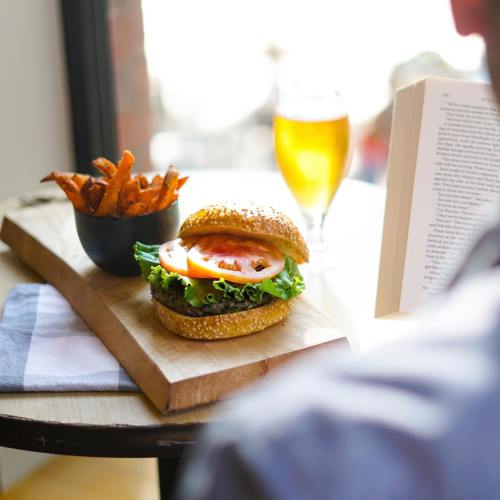 A burger with lettuce and tomato, sweet potato fries, and a glass of beer on a table next to an open book.