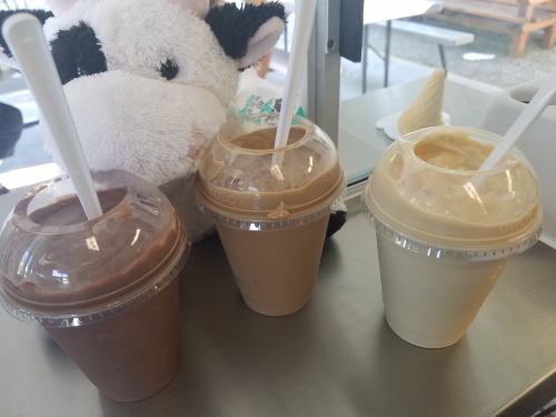 Three milkshakes in clear cups—chocolate, vanilla, and a light brown flavor—next to a plush cow toy.