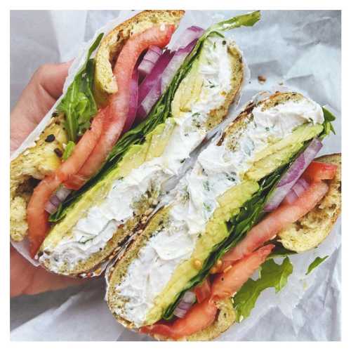 A hand holding a freshly cut sandwich filled with cream cheese, avocado, lettuce, tomato, and red onion on multigrain bread.