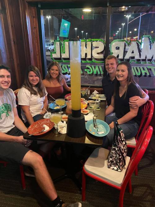 A group of five friends enjoying a meal at a restaurant, with drinks and food on the table. Brightly lit atmosphere.