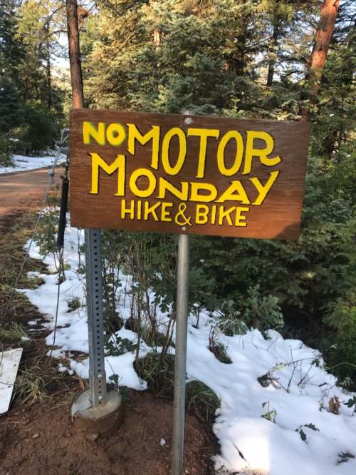 Wooden sign reading "No Motor Monday Hike & Bike" in a forested area with some snow on the ground.