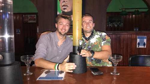 Two smiling men sit at a bar, posing with a large yellow pole between them, drinks and a menu on the table.