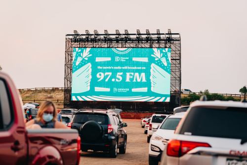 A large outdoor screen displays "The world's music will broadcast on 97.5 FM" at a drive-in event, with cars parked nearby.