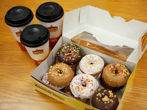 A box of assorted donuts with sprinkles and a chocolate coating, alongside three cups of coffee on a wooden table.