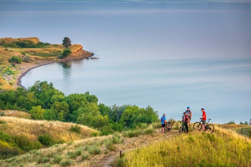 Four cyclists stand on a grassy hill overlooking a calm lake and lush greenery along the shoreline.