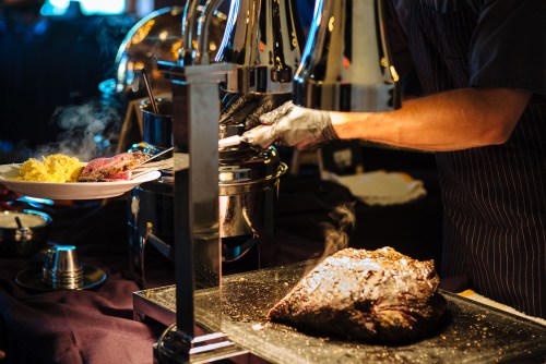 A chef serves sliced meat from a carving station, with a plate of food and steam rising in a dimly lit setting.