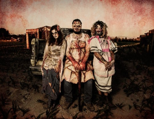 Three horror-themed characters in tattered costumes, covered in fake blood, pose ominously in a desolate outdoor setting.