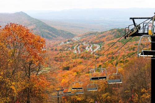A scenic view of autumn foliage from a ski lift, showcasing vibrant orange and yellow trees and winding roads below.