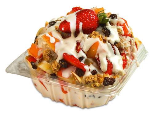 A colorful fruit salad topped with yogurt, granola, and a strawberry, served in a clear plastic container.