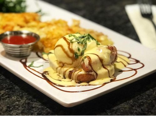 A plate of eggs Benedict drizzled with sauce, served with hash browns and a side of ketchup.