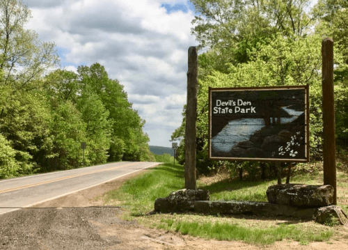 Sign for Devil's Den State Park beside a road, surrounded by lush green trees and a cloudy sky.