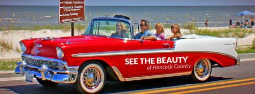 A classic red and white convertible drives along a coastal road with a beach in the background, promoting Hancock County.