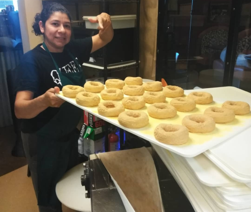 A person holds a tray of freshly made doughnuts in a kitchen setting, with several doughnuts arranged neatly.