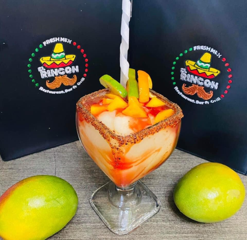 A colorful fruit cocktail in a glass, garnished with mango and green fruit, with two whole mangoes beside it.