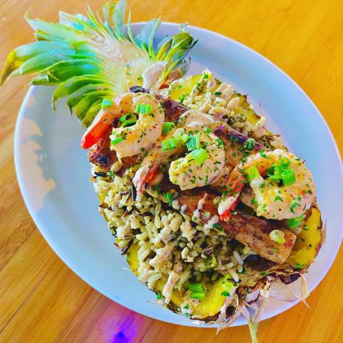 A pineapple half filled with rice, grilled shrimp, and garnished with green onions and herbs on a white plate.