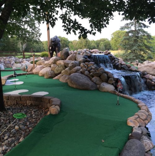 A mini-golf course featuring a rocky waterfall, a bear statue, and lush greenery in the background.