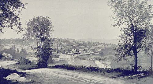 A vintage landscape showing a small town with houses, trees, and a winding road, set against a hilly backdrop.
