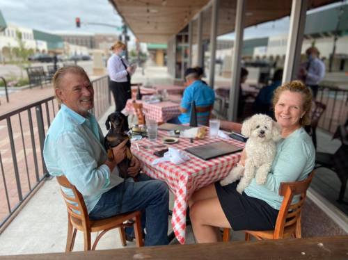 A man and woman sit at a restaurant table with their dogs, enjoying a meal outdoors. Red and white checkered tablecloth.
