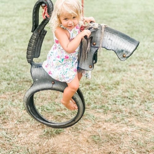 A young child in a floral dress plays on a tire swing shaped like a horse in a grassy area.