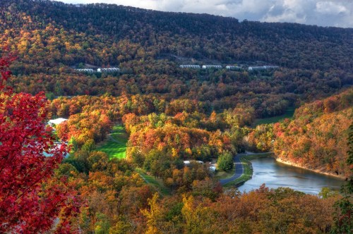 A vibrant autumn landscape featuring colorful trees, a winding river, and rolling hills under a cloudy sky.