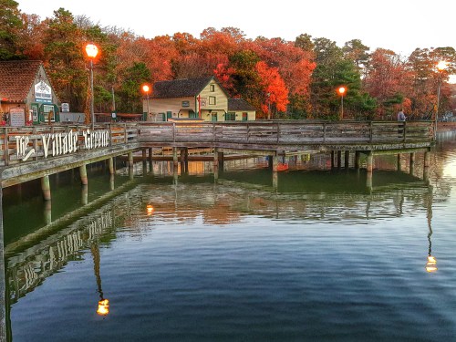 A wooden pier extends over calm water, reflecting autumn trees and a quaint building, with warm lights glowing at dusk.