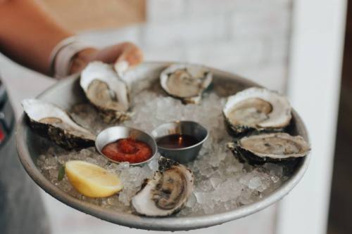 A metal platter holds fresh oysters on ice, accompanied by lemon wedges and dipping sauces.