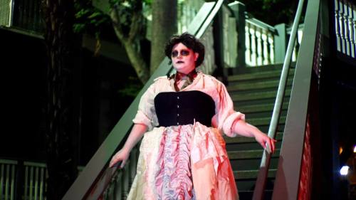 A person in a dramatic costume with dark makeup stands on a staircase, surrounded by trees and dim lighting.