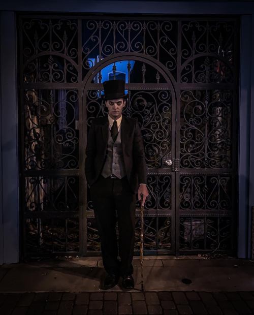 A man in a top hat and suit stands with a cane in front of an ornate wrought iron gate at night.