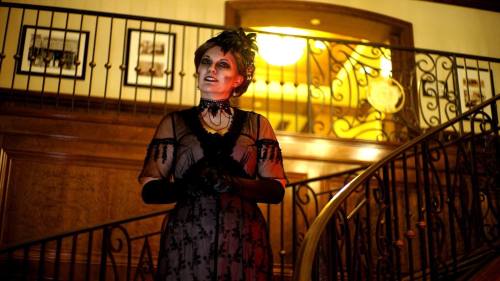 A woman in a vintage black dress stands on a staircase, illuminated by warm light, with a dramatic expression.