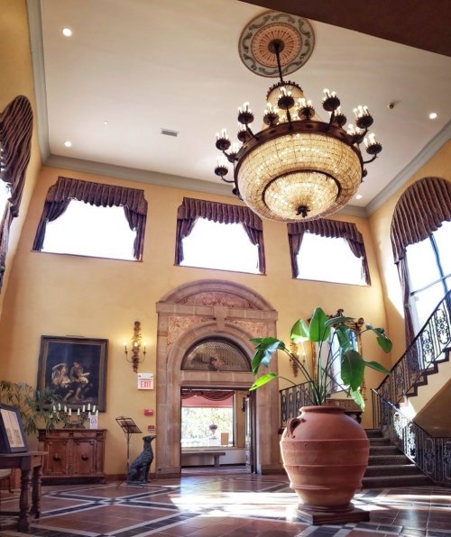 Elegant lobby with a large chandelier, decorative windows, and a potted plant, featuring ornate architecture and warm lighting.