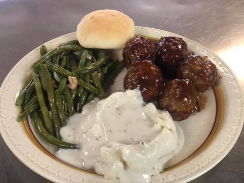 A plate of meatballs with barbecue sauce, green beans, mashed potatoes with gravy, and a dinner roll.
