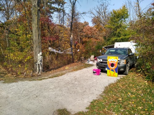 A spooky outdoor scene with a skeleton, colorful decorations, and a truck surrounded by autumn foliage.