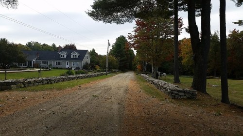 A gravel road lined with stone walls, leading to a house surrounded by trees with autumn foliage.