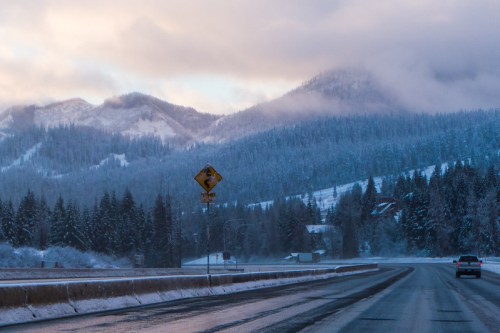 A snowy mountain landscape with a winding road, trees, and a caution sign under a cloudy sky.