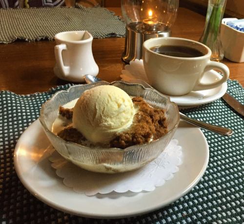A bowl of dessert topped with vanilla ice cream, accompanied by a cup of coffee and a small creamer on a table.