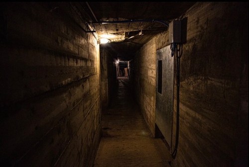 Dimly lit underground corridor with concrete walls and a barred exit in the distance.