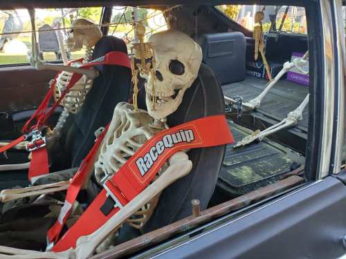 Two skeletons in racing seats, secured with red harnesses, inside a car.