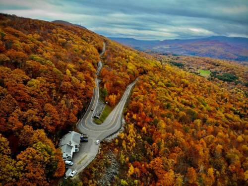 Aerial view of a winding road through vibrant autumn foliage in the mountains, with a small building nearby.