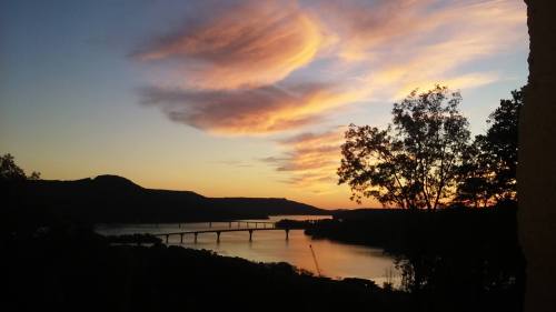 Sunset over a river with a bridge, framed by trees and mountains, showcasing vibrant clouds in the sky.
